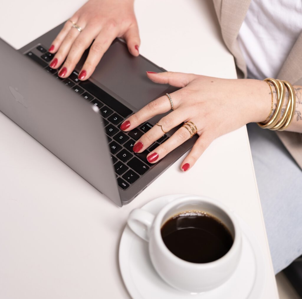 Frauenhände tippend am Laptop. Daneben steht eine Tasse mit schwarzem Kaffee.