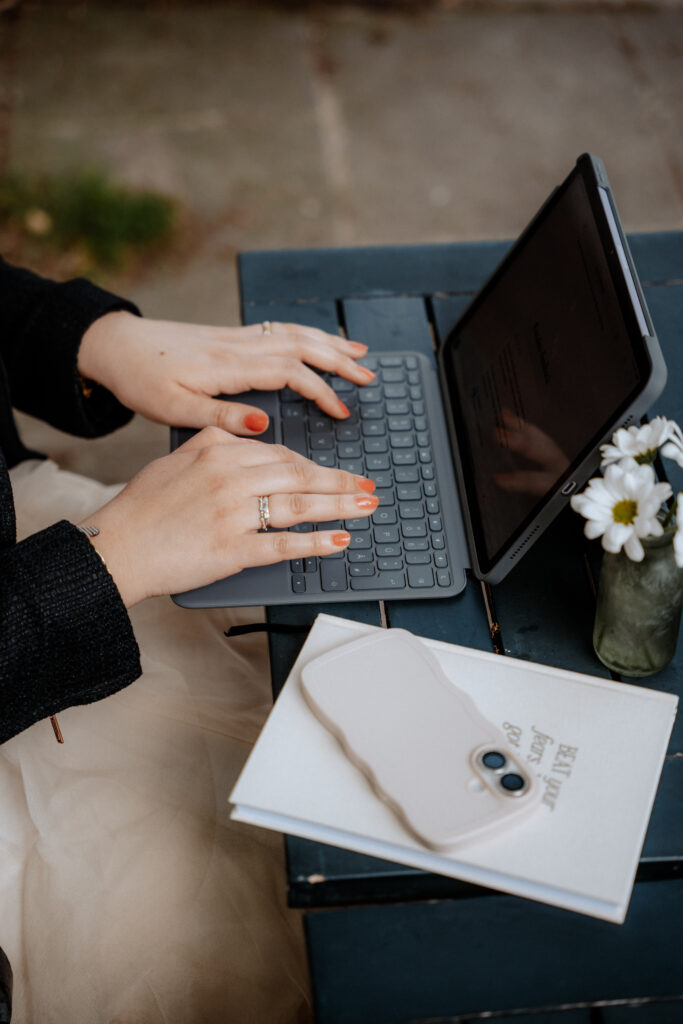Nadine von Content Instinct tippt auf der Tastatur eines Laptops. Zu sehen sind nur ihre Finger. Daneben ein Notizbuch und ein Handy.